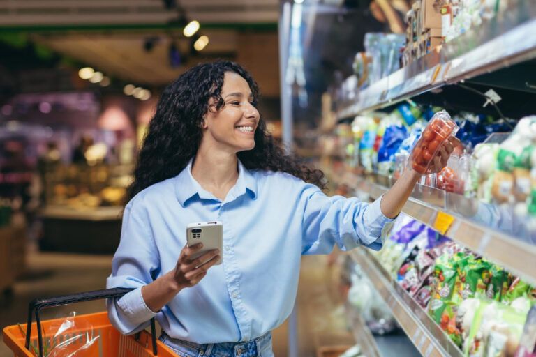 Person holding a smartphone and a shopping basket, smiling while selecting packaged food from a grocery store shelf.