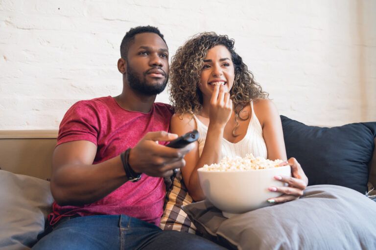 A man and woman sit on a couch watching TV, with a remote in the man's hand and a bowl of popcorn in the woman's lap.