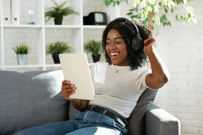 A person wearing headphones sits on a couch, smiling and looking at a tablet. Shelves with plants are in the background.