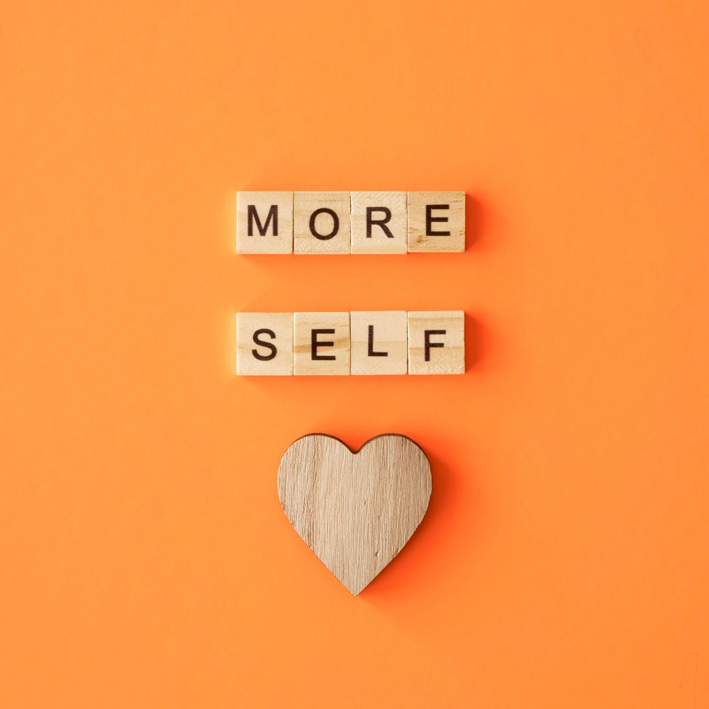 Wooden letter tiles spell "MORE SELF" above a wooden heart on an orange background.