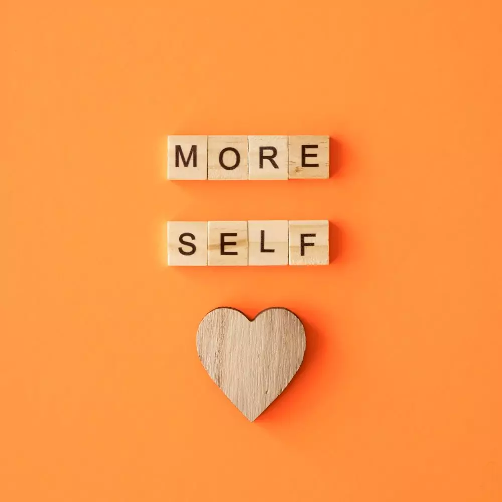 Wooden letter tiles spell "MORE SELF" above a wooden heart on an orange background.