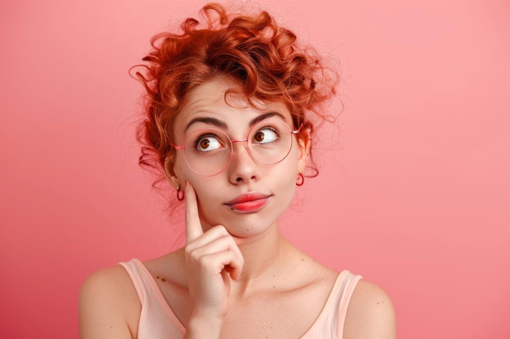 A person with curly red hair and pink glasses thoughtfully touching their cheek against a pink background.