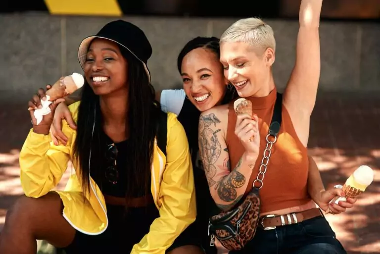 Three people sitting and enjoying ice cream cones while smiling. One person wears a yellow jacket and bucket hat, another with braided hair, and a third with short hair and tattoos.