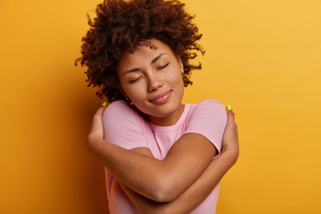 A person with curly hair in a pink shirt hugs themselves with a content expression against a yellow background.