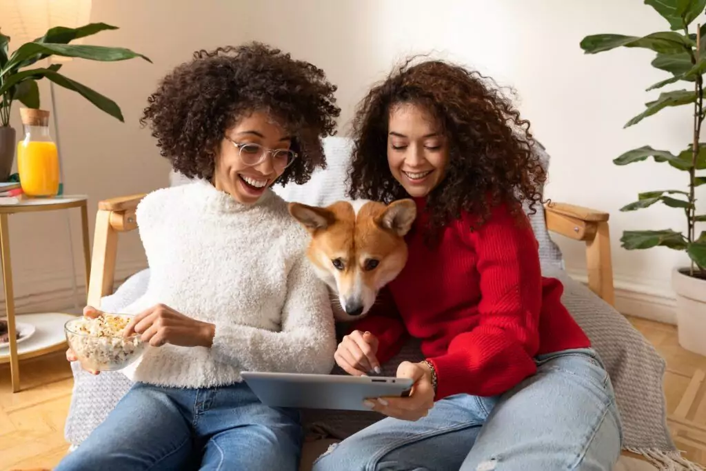 Two people sitting on a couch with a dog. One holds popcorn, the other uses a tablet.